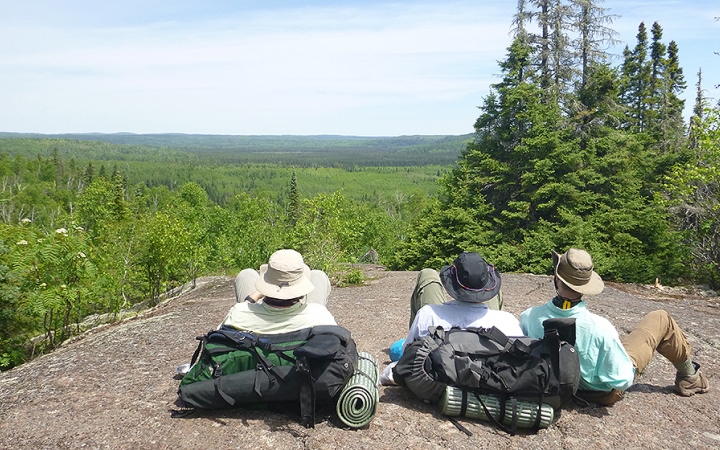 Three people sit on a rocky overlook and lean back on their backpacks, taking in the vast green forrest below.